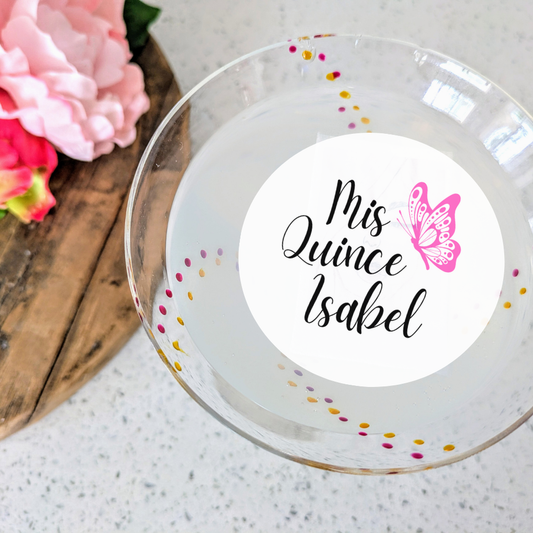 A personalized edible drink topper with the text 'Mis Quince Isabel' printed on it, featuring a butterfly design, placed on a glass.