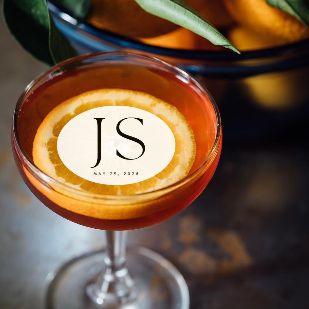 Cocktail with an orange slice and custom wedding cocktail topper 'JS' logo, blurred oranges in the background