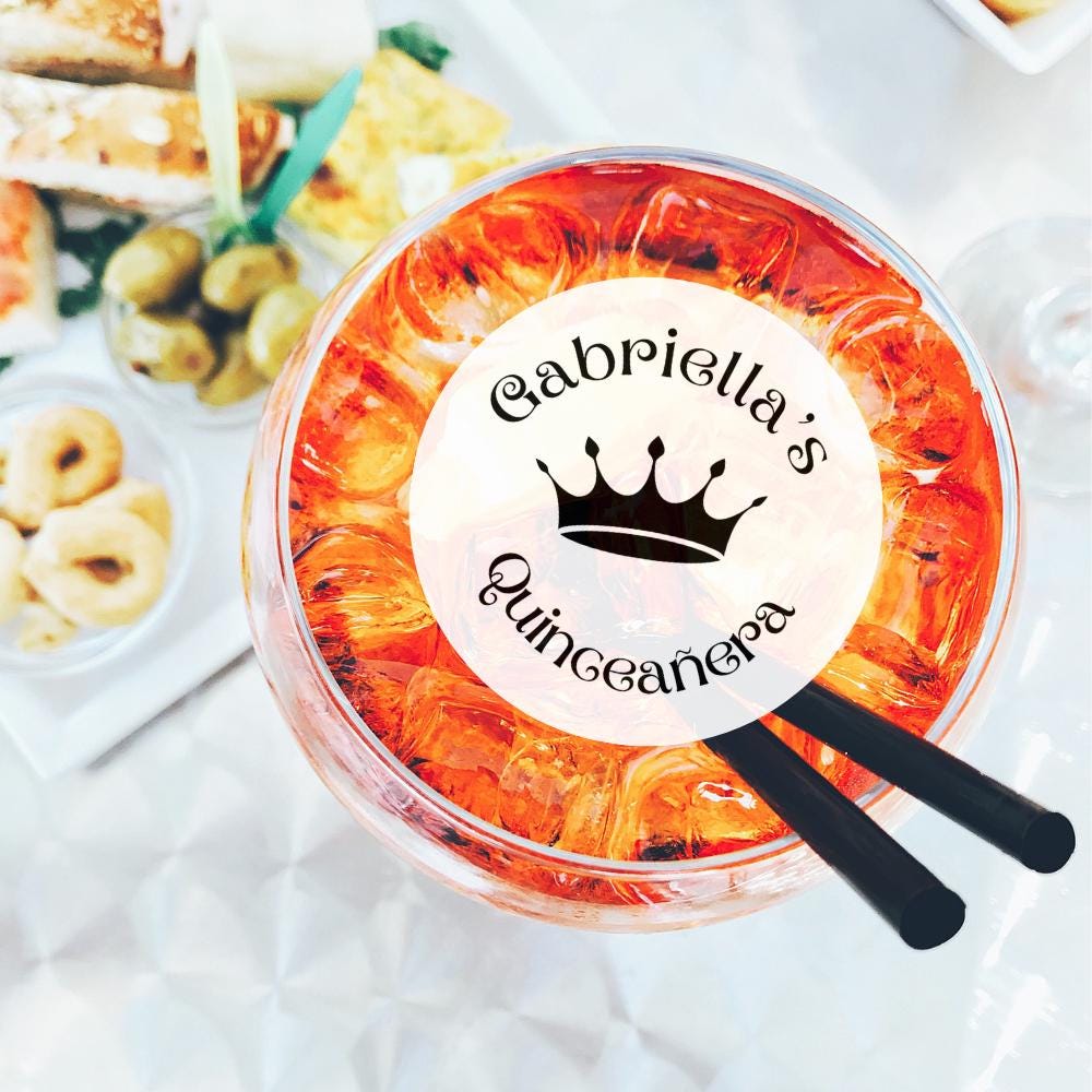 Clear plastic cup with orange design and 'Gabriella's Quinceanera' text, surrounded by snacks on a white surface.