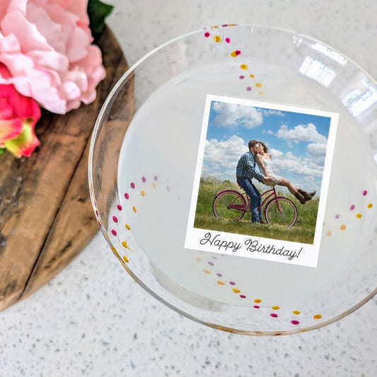 Glass with colorful polka dots featuring a edible image drink topper. Photo is of a couple on a bicycle and 'Happy Birthday!' text, placed on a wooden surface with flowers.