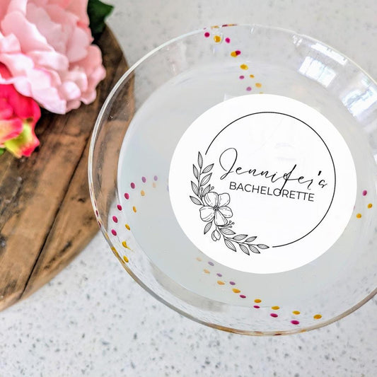 Glass plate with a personalized edible drink topper for a bachelorette party, surrounded by flowers on a wooden board.