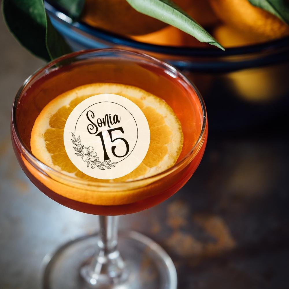 Cocktail garnish with an orange slice and 'Sonia 15' edible topper, blurred oranges in the background