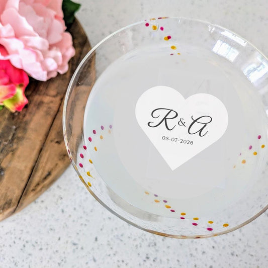 Glass plate with a heart-shaped edible cocktail garnish topper of initials and date, surrounded by flowers on a wooden surface.