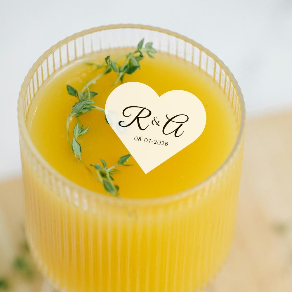 Glass of yellow liquid with a heart-shaped edible sticker featuring initials and date, garnished with a sprig of thyme.