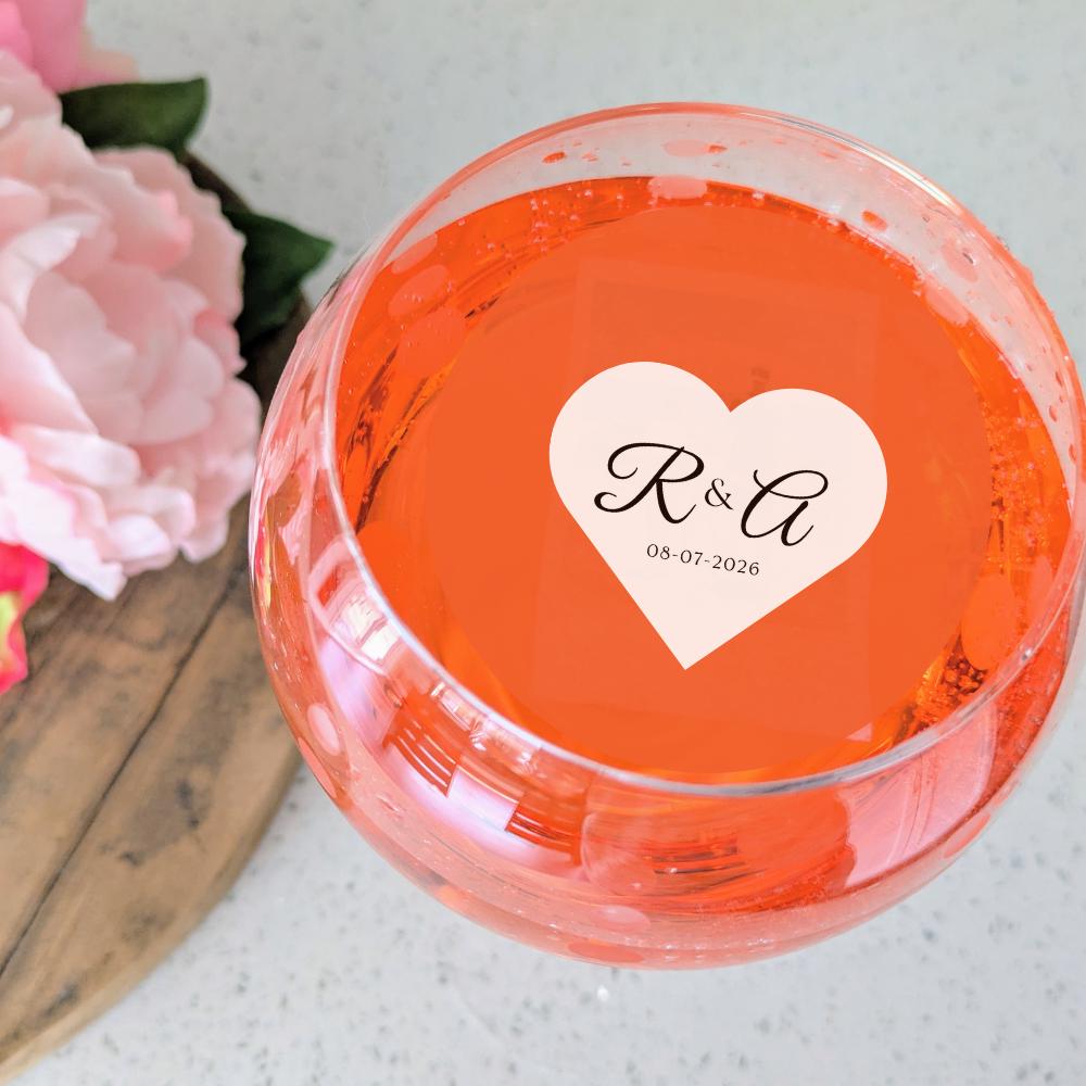 Clear glass bowl with a custom  heart-shaped edible drink sticker featuring initials and date, surrounded by pink flowers.