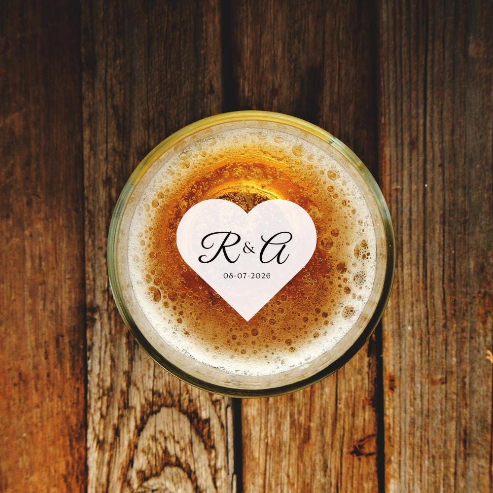Glass of beer with a heart-shaped beverage topper featuring initials and date on a wooden surface