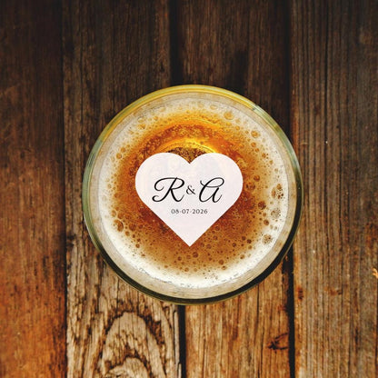 Glass of beer with a heart-shaped beverage topper featuring initials and date on a wooden surface